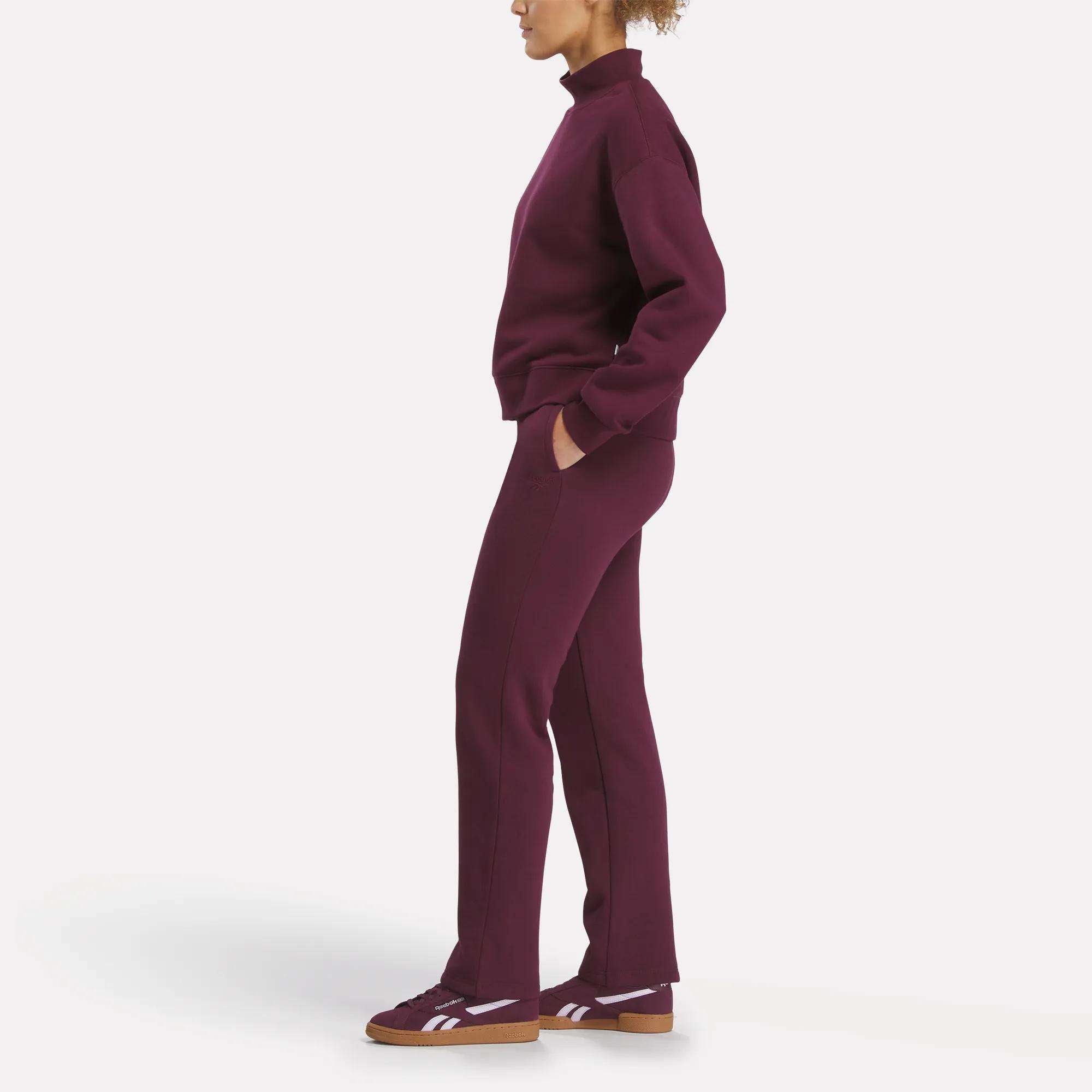 A woman stands in profile wearing Reebok's Women's Cozy Classic Sweatpants in burgundy, styled with a matching high-neck top and burgundy sneakers with white stripes and brown soles, set against a plain white background.