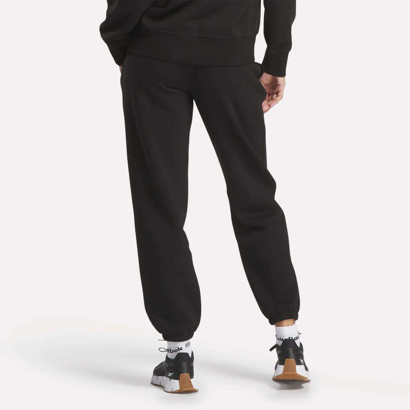 A person stands with one hand in their pocket, viewed from behind against a plain white background, wearing Reebok Women's Medium Vector Sweatpants and a black sweatshirt, paired with Reebok sneakers and white socks.