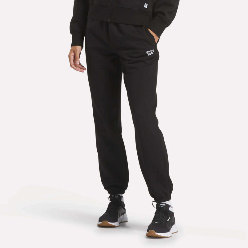 A person models Reebok Women's Medium Vector Sweatpants in black with a white logo, paired with a black hoodie, black athletic shoes, and white socks against a plain white background. The person's head is not visible.