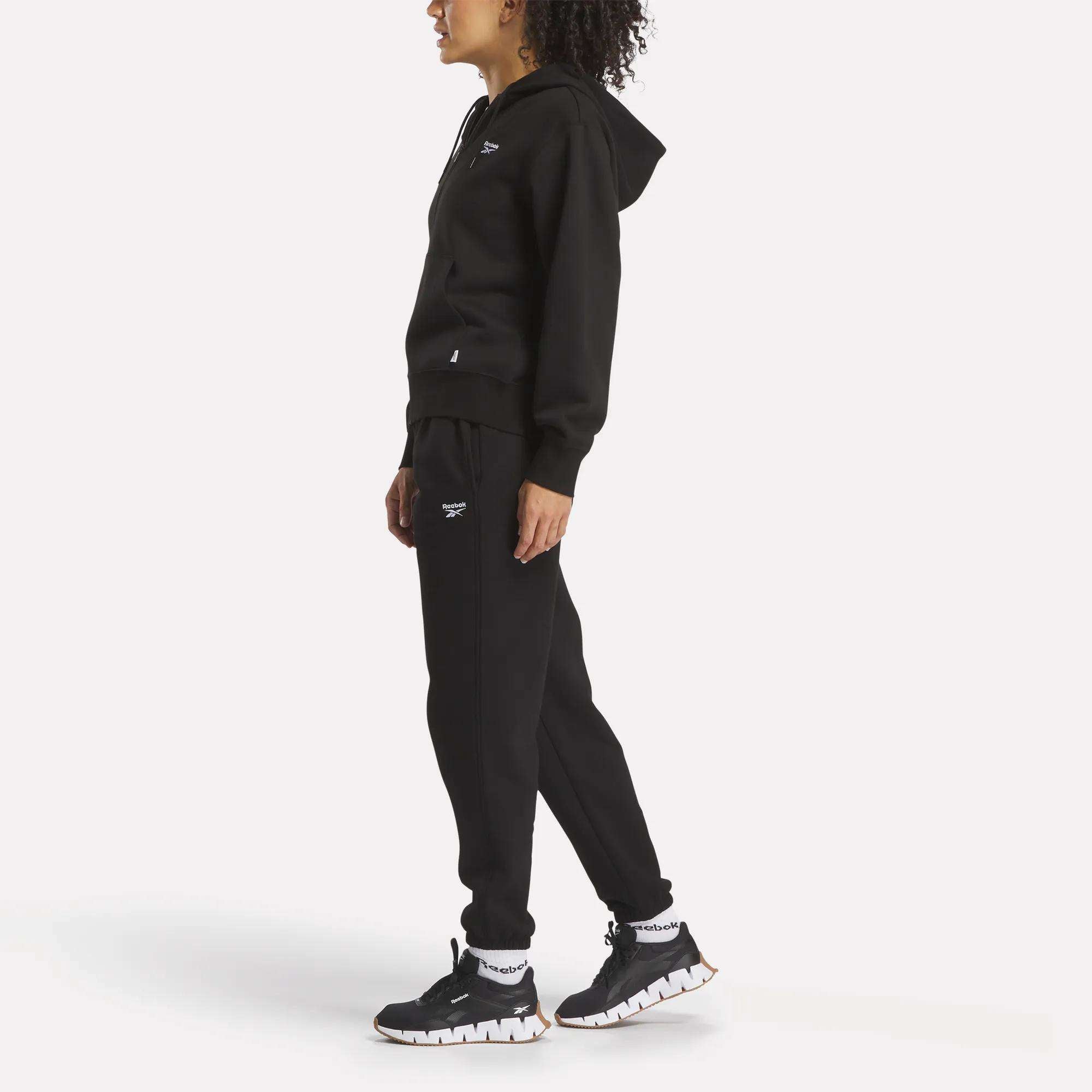 A person in profile wears Reebok Women's Medium Vector Sweatpants featuring a small white logo, paired with a black hooded sweatshirt and black sneakers with white soles, all shown against a plain white background.