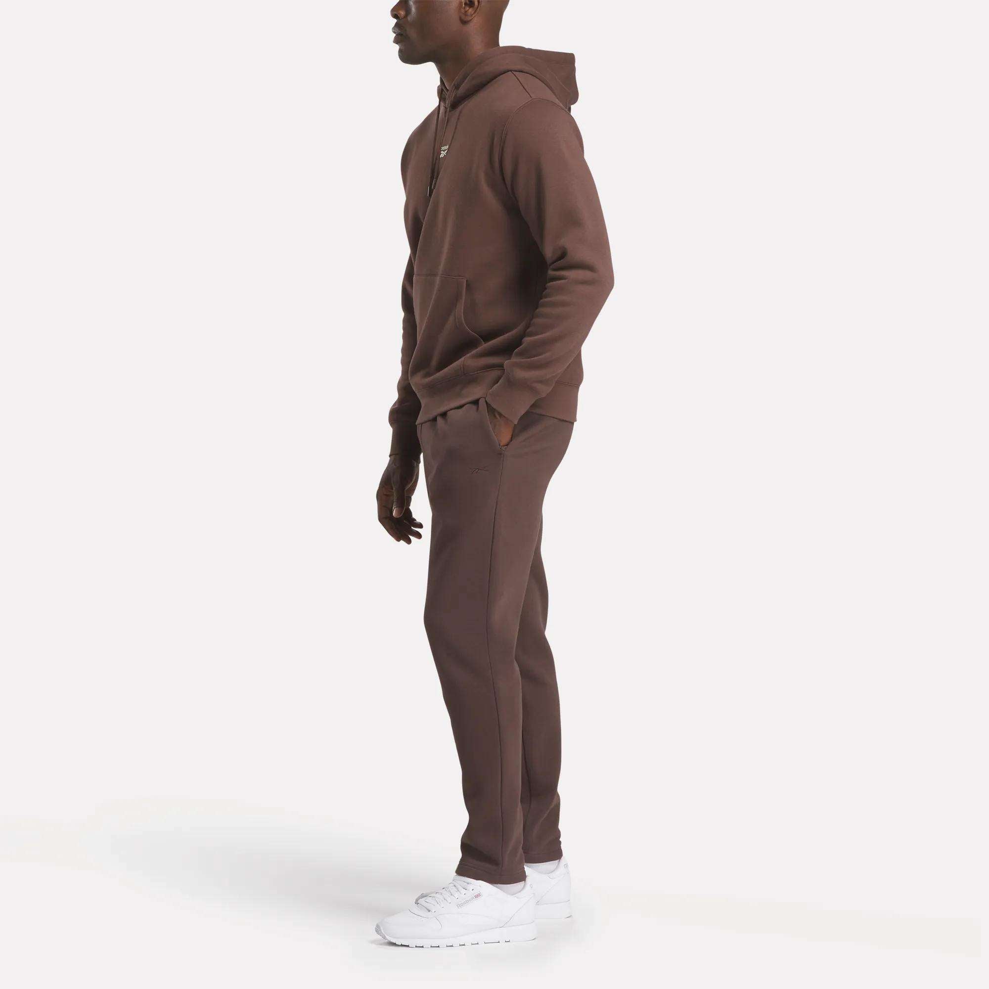 A person stands sideways in a Reebok Men's Finisher Hoodie and matching sweatpants with white sneakers, set against a plain white background.