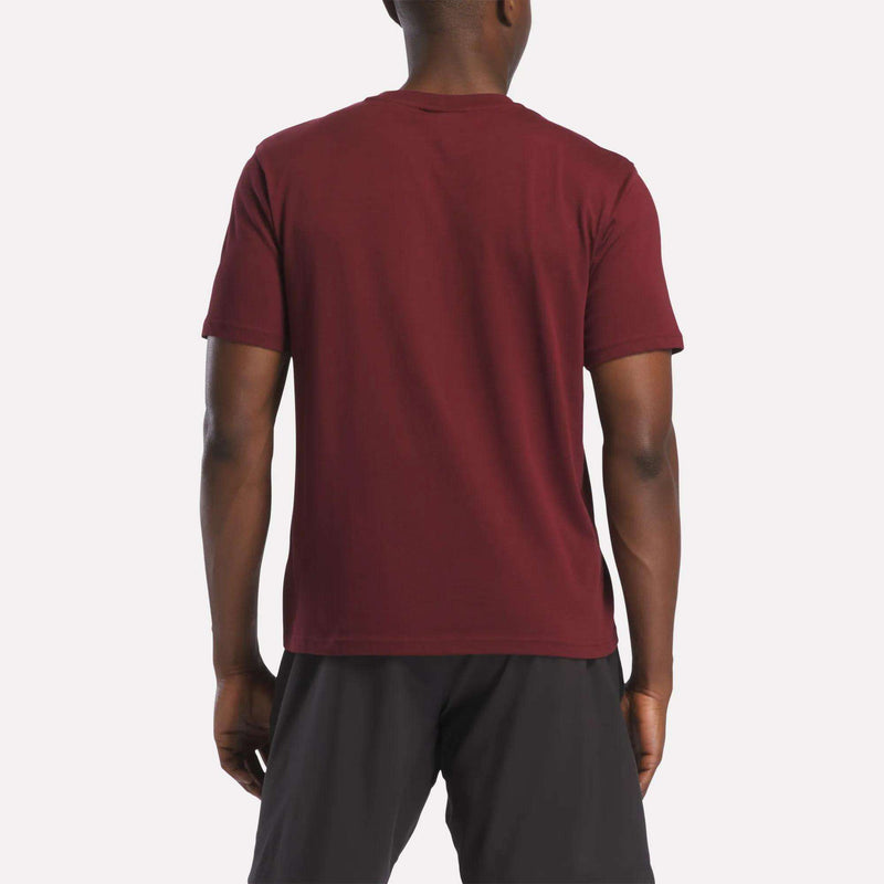 A person stands with their back to the camera, wearing a Reebok Men's Logo Crewneck T-Shirt in maroon and black shorts against a light background.