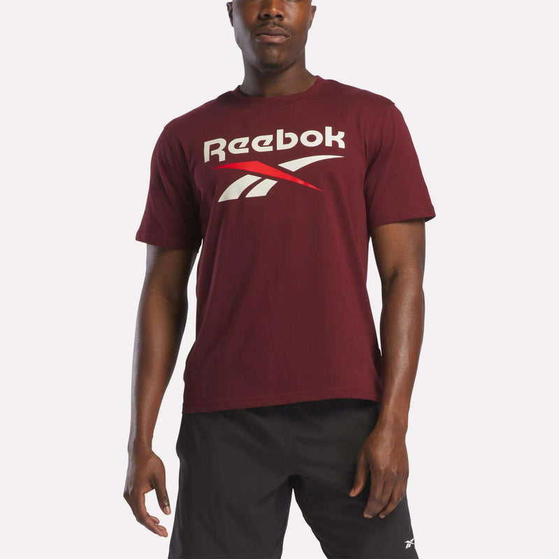 A man stands against a plain white background wearing the Reebok Men's Logo Crewneck T-Shirt in maroon with a large white and red vector logo, paired with black shorts.