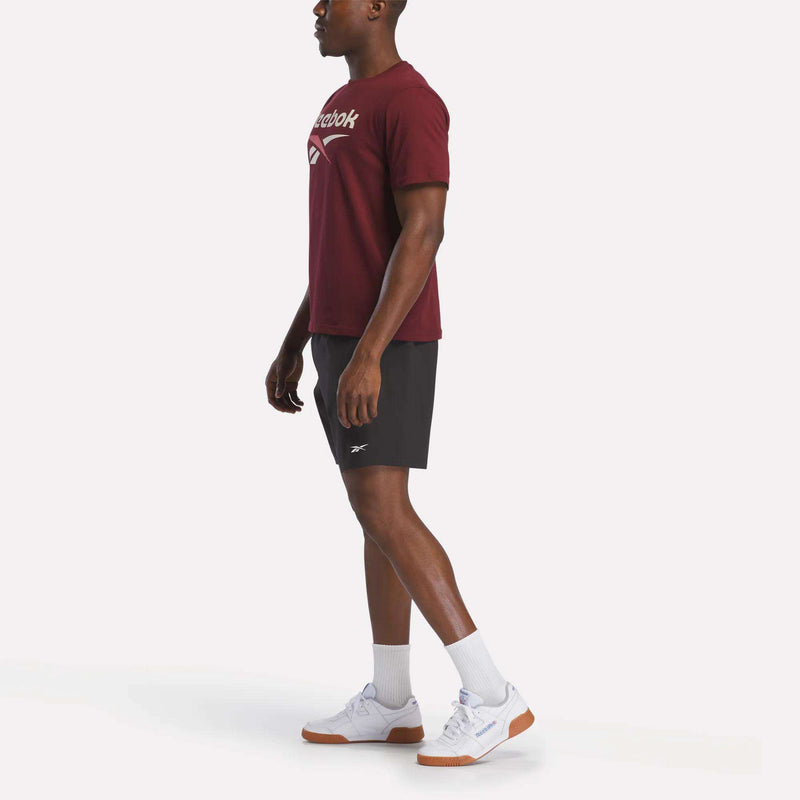 A man wears a Reebok Men's Logo Crewneck T-Shirt in maroon, paired with black shorts, white crew socks, and white sneakers with gum soles, standing against a plain white background.