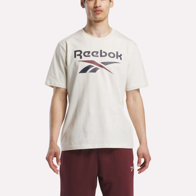A person wearing a white Reebok Men's Logo Crewneck T-Shirt with a bold front logo and maroon shorts, standing against a plain white background.