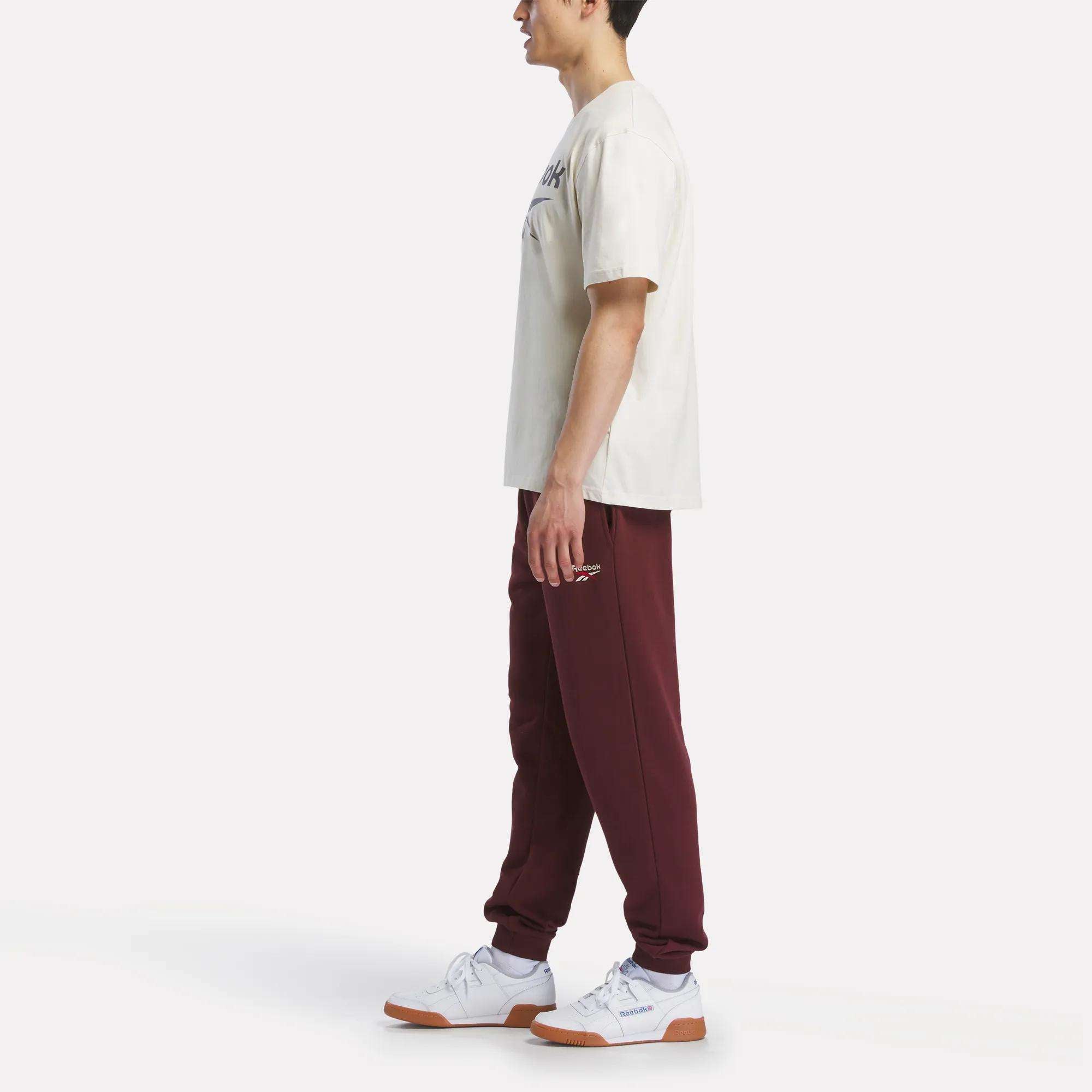 A person stands in profile, wearing a cream Reebok Men's Logo Crewneck T-Shirt, maroon sweatpants, and white sneakers with brown soles against a plain white background.