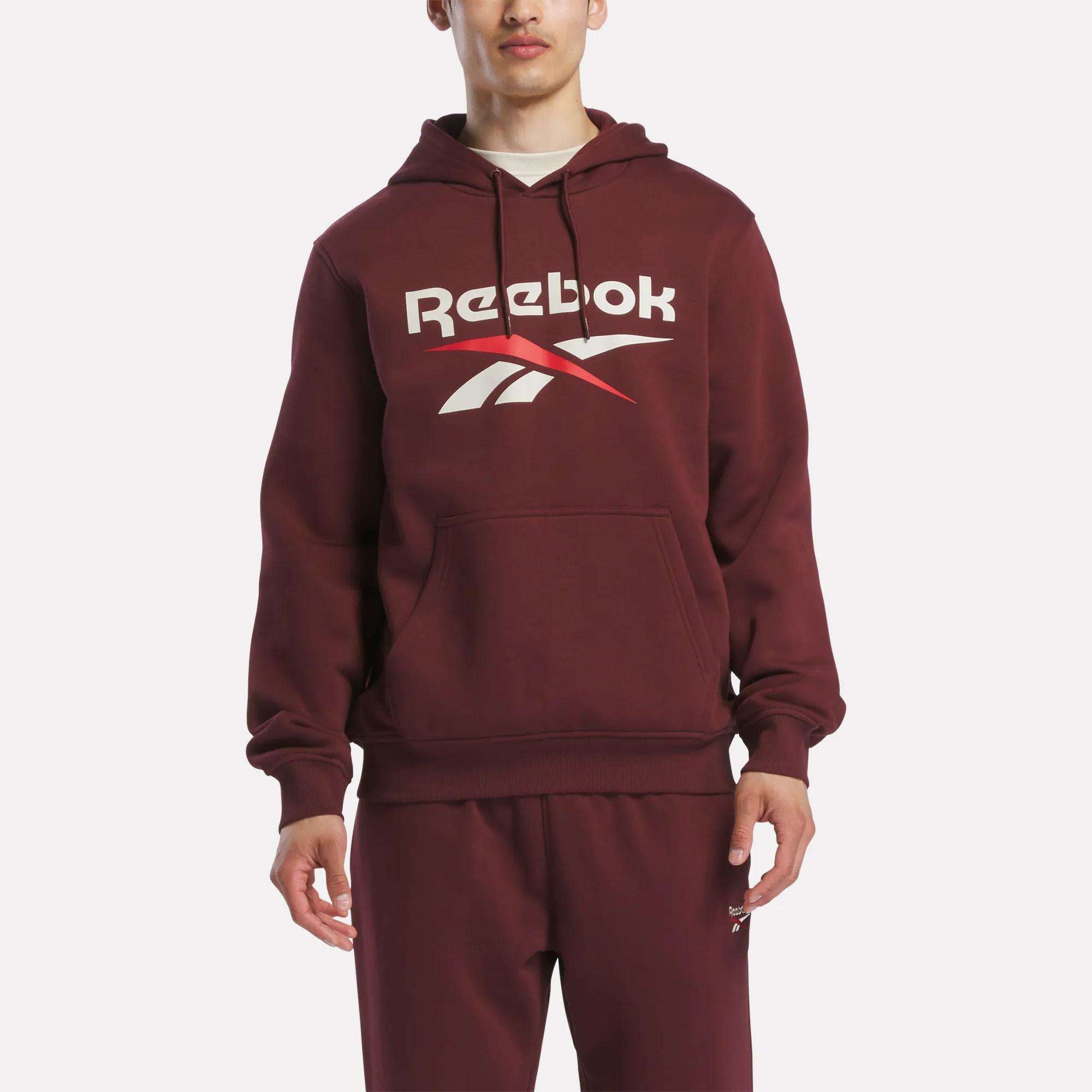 A person stands facing forward in a maroon Reebok Men's Logo Hoodie, featuring a bold chest logo and made from soft cotton-blend fleece for comfort. Matching sweatpants complete the look.