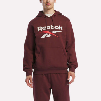 A person stands facing forward in a maroon Reebok Men's Logo Hoodie, featuring a bold chest logo and made from soft cotton-blend fleece for comfort. Matching sweatpants complete the look.