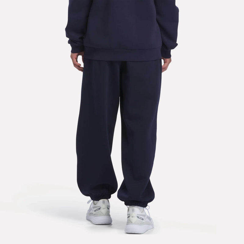 A person is shown from the waist down, facing away from the camera, wearing Reebok Men’s Logo Relaxed Fleece Pants in navy blue paired with a matching sweatshirt and white sneakers, standing against a plain white background.