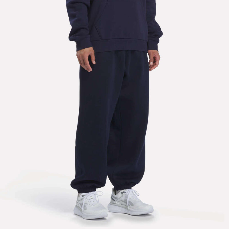 A person is standing with their hands by their sides, wearing Reebok Men’s Logo Relaxed Fleece Pants in Vector Navy along with a navy blue sweatshirt. They are also wearing white athletic shoes. The background is plain and light-colored.