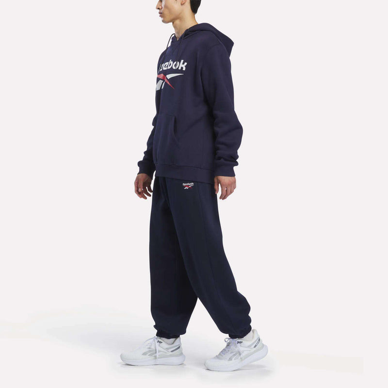 A person is wearing a navy blue Reebok hoodie and Men’s Logo Relaxed Fleece Pants by Reebok, paired with white sneakers, standing sideways against a plain white background.