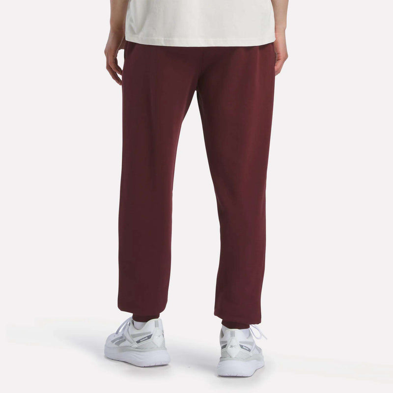 A person stands facing away against a plain light backdrop, showing only their lower half in maroon Reebok Men's French Terry Logo Joggers, paired with a white shirt and white sneakers.