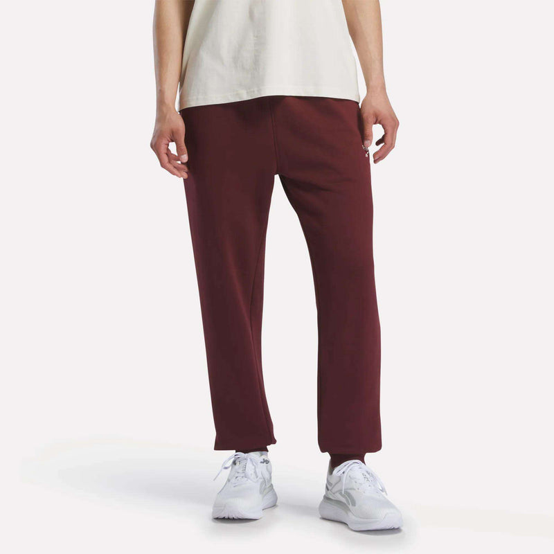 A person models Reebok Men's French Terry Logo Joggers in burgundy, paired with a white T-shirt and white sneakers, against a plain white background. The image is cropped from the shoulders down.