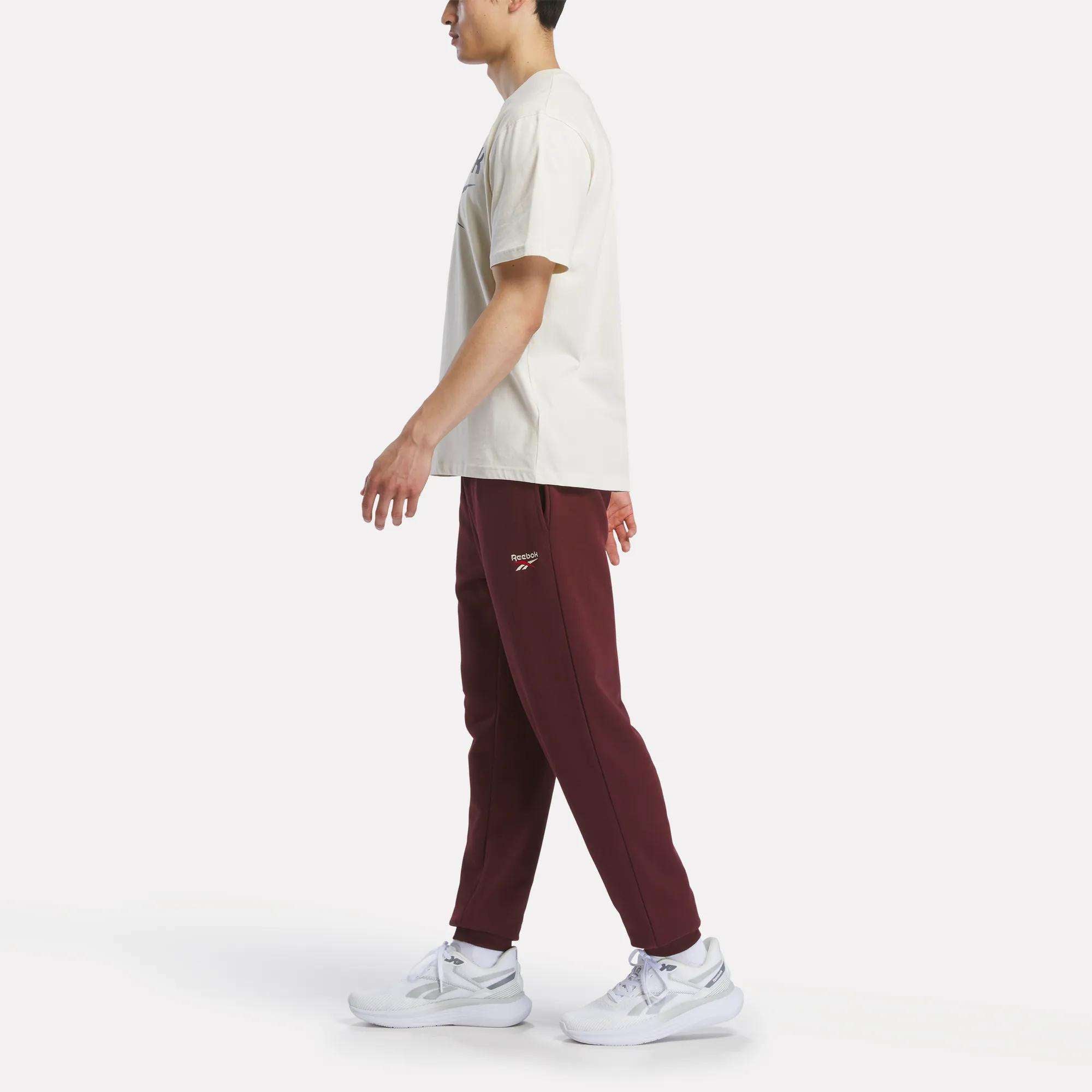A person in a beige t-shirt, Reebok Men's French Terry Logo Joggers, and white sneakers walks left against a plain white background.