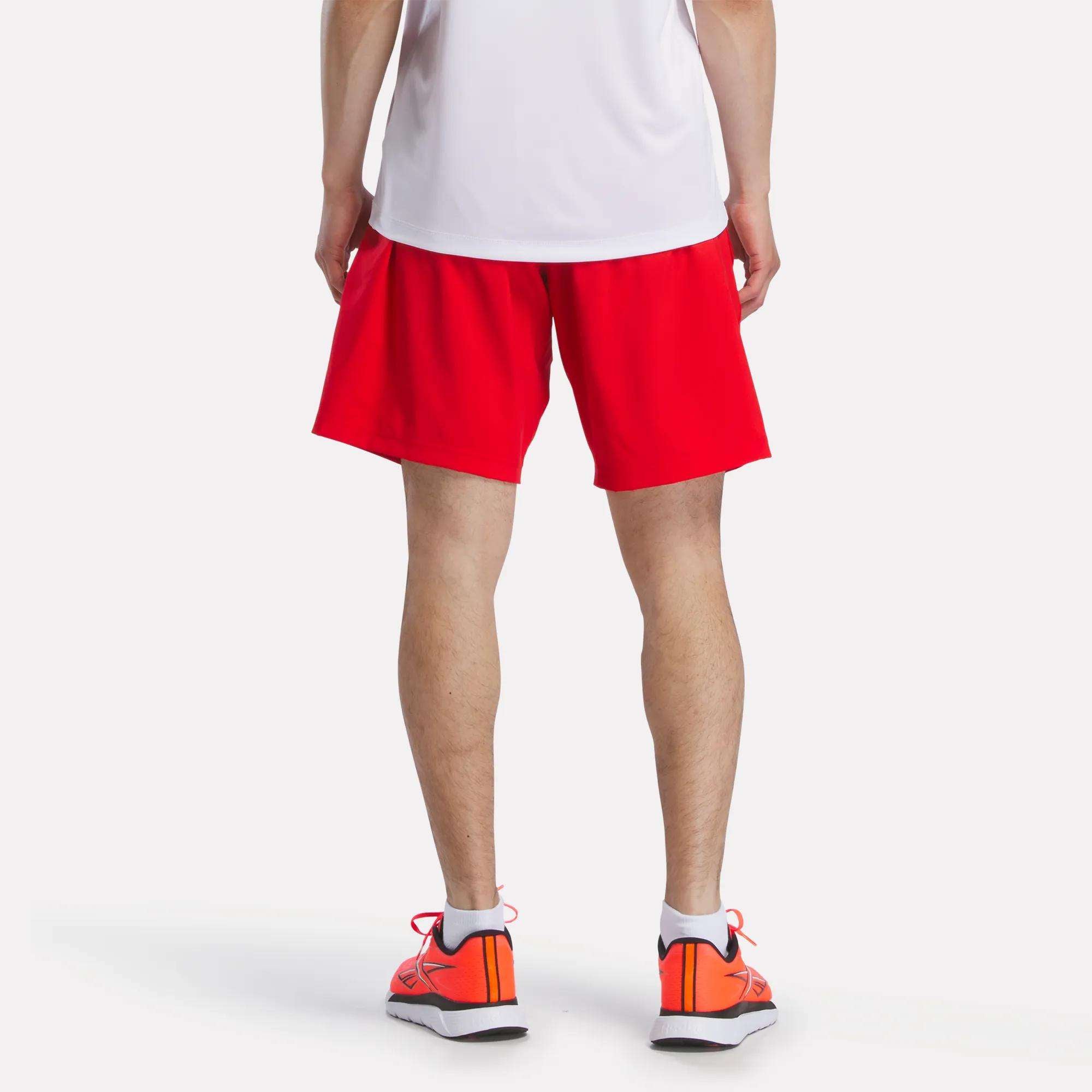 A man stands facing away from the camera, wearing a white shirt and bright red Reebok Men's Woven Shorts. He also sports orange running shoes with white soles against a plain white background.