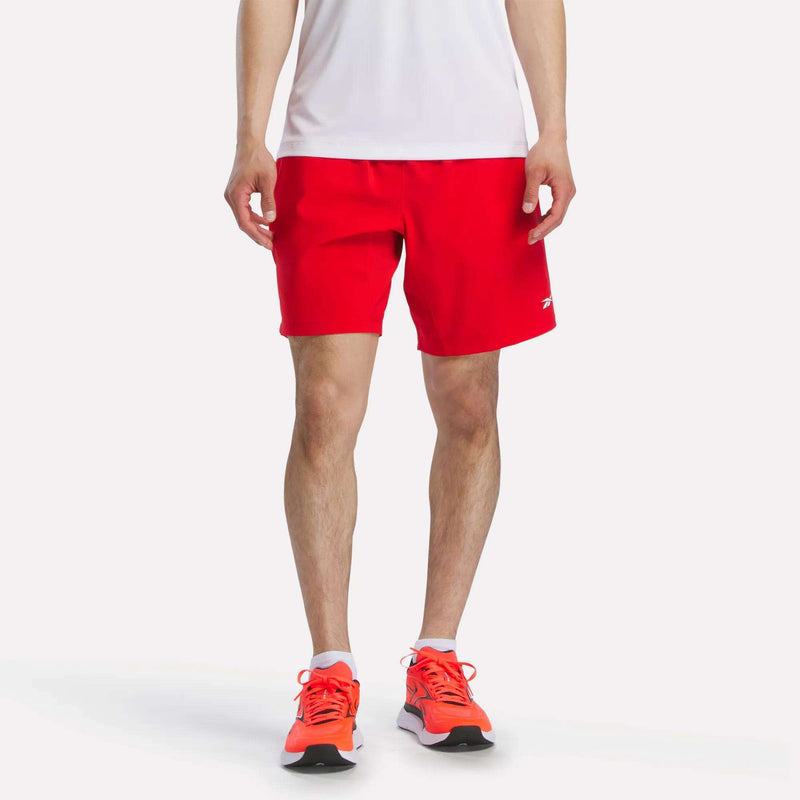 A man models Reebok Men's Woven Shorts in bright red, paired with red running shoes and a white shirt. The photo shows only his lower torso and legs facing forward against a plain white background.