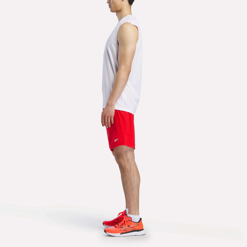 A man stands in profile wearing a white sleeveless shirt, Reebok Men's Woven Shorts, and bright orange athletic shoes against a plain white background.