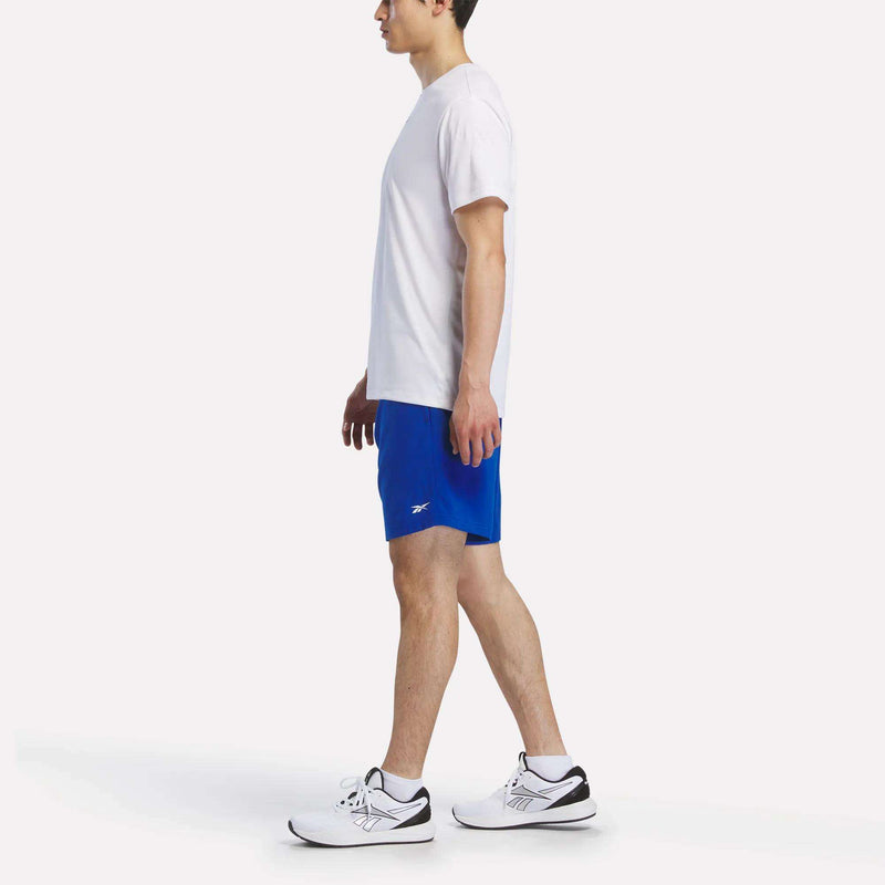 A man in profile wears a white T-shirt, Reebok Men's Woven Shorts in bright blue, and white sneakers with black details, set against a plain white background.