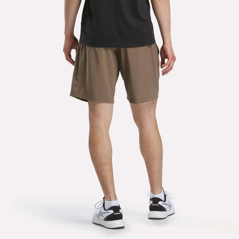 A person in a black shirt, Reebok Men's Woven Shorts in light brown, and white sneakers stands with their back to the camera against a plain white background.