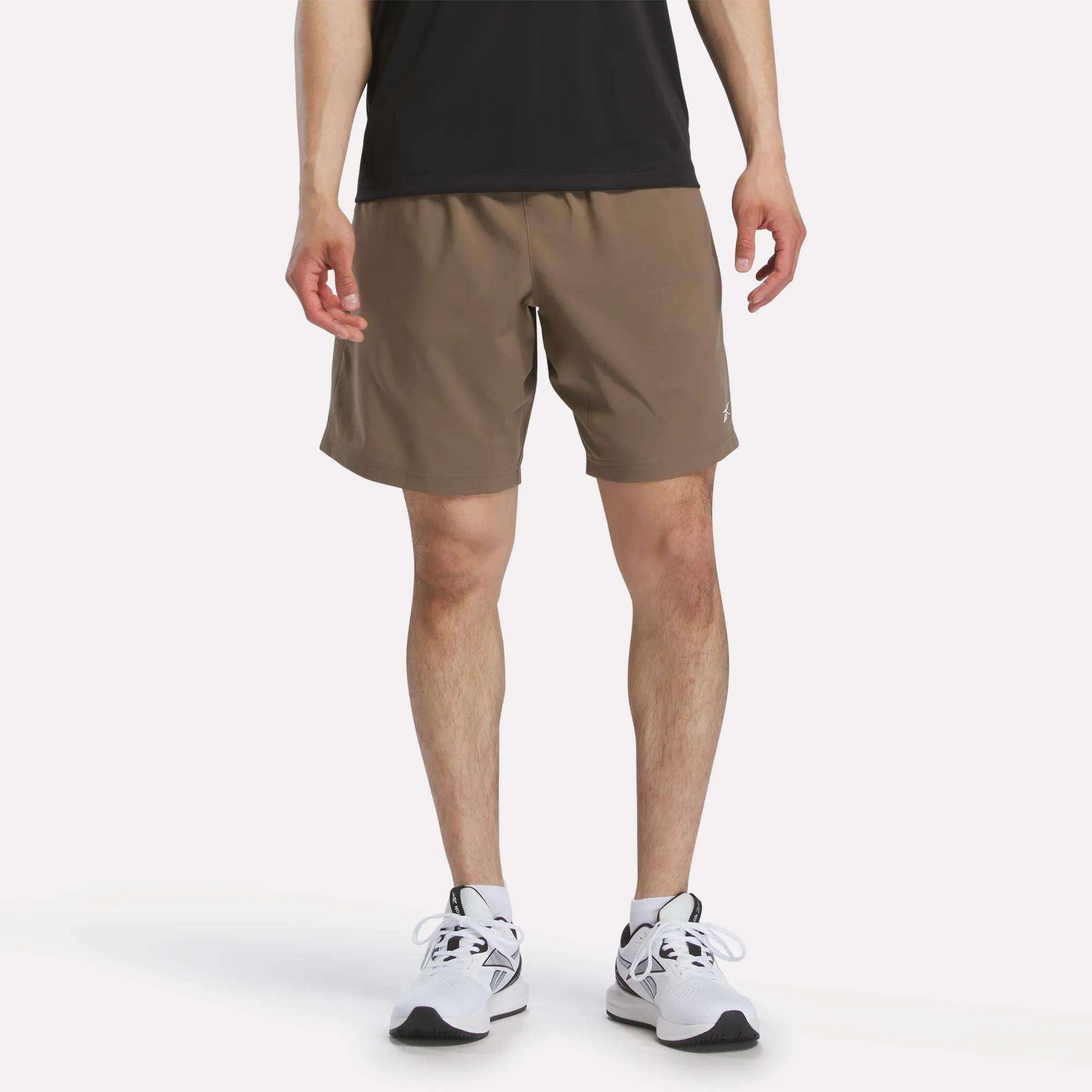 A person wearing Reebok Men's Woven Shorts stands on a plain white background, shown from the shoulders down, paired with a black shirt and white running shoes.