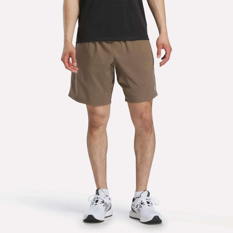 A person wearing Reebok Men's Woven Shorts stands on a plain white background, shown from the shoulders down, paired with a black shirt and white running shoes.