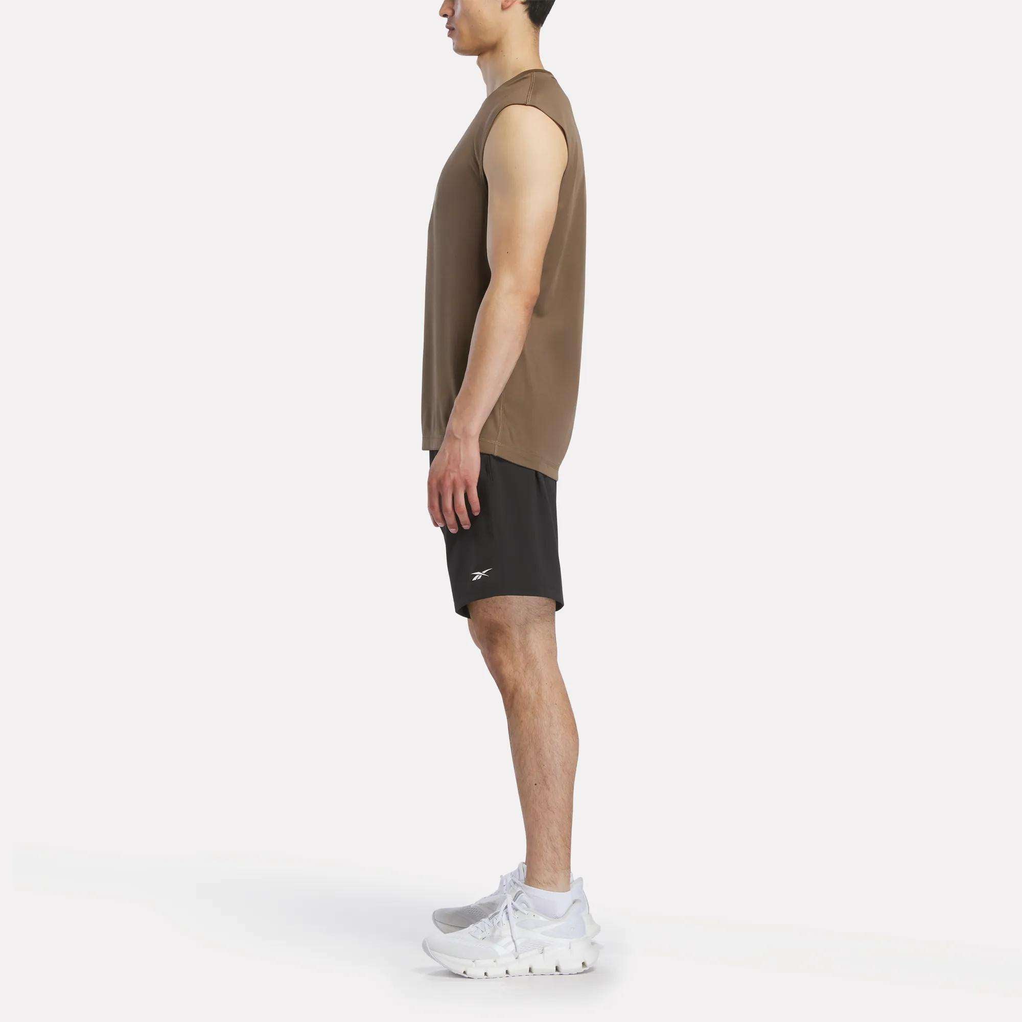 A man stands in profile facing left, wearing a Reebok Men's Sleeveless Tech Training T-Shirt in brown, black shorts, and white athletic shoes against a plain white background.