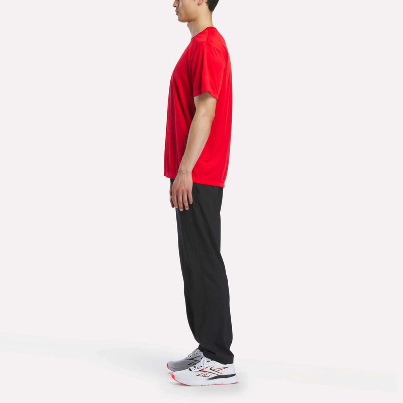 A person stands in profile facing left, wearing a red Reebok Men's Short Sleeve Tech Training T-Shirt, black pants, and white athletic shoes against a plain white background.