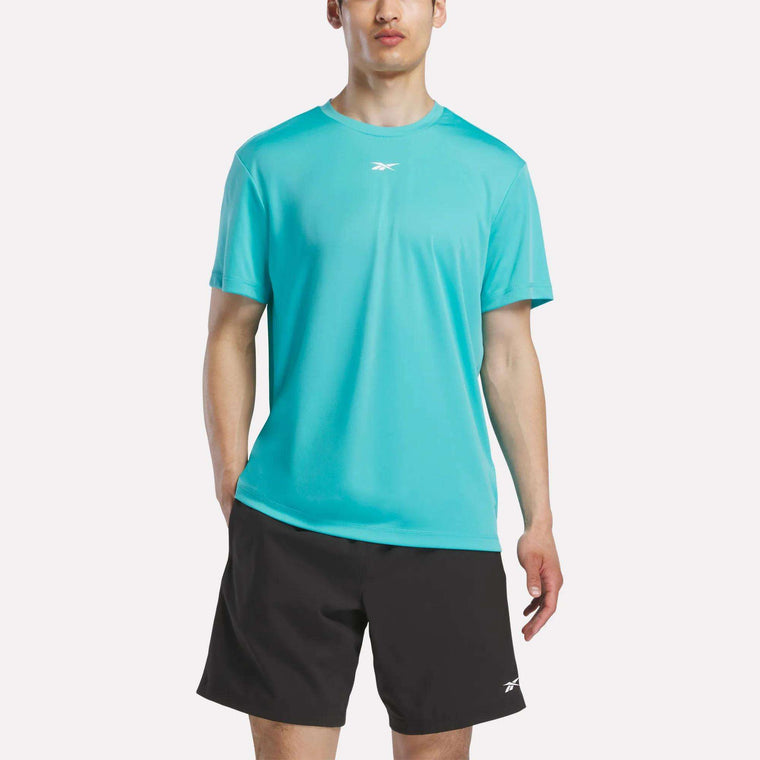 Against a plain light background, a person wears Reebok’s Men’s Short Sleeve Tech Training T-Shirt in turquoise with sweat-wicking fabric and black shorts; both display small white Reebok logos.