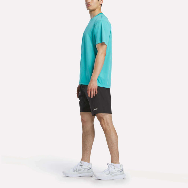 A man stands sideways in a Reebok Men's Short Sleeve Tech Training T-Shirt (turquoise), black Reebok shorts, and white sneakers against a plain white background. The gym shirt features sweat-wicking fabric for optimal comfort.