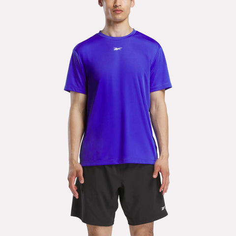 A man in a bright blue Reebok Men's Short Sleeve Tech Training T-Shirt stands facing forward against a plain light background, showcasing this sweat-wicking gym shirt.