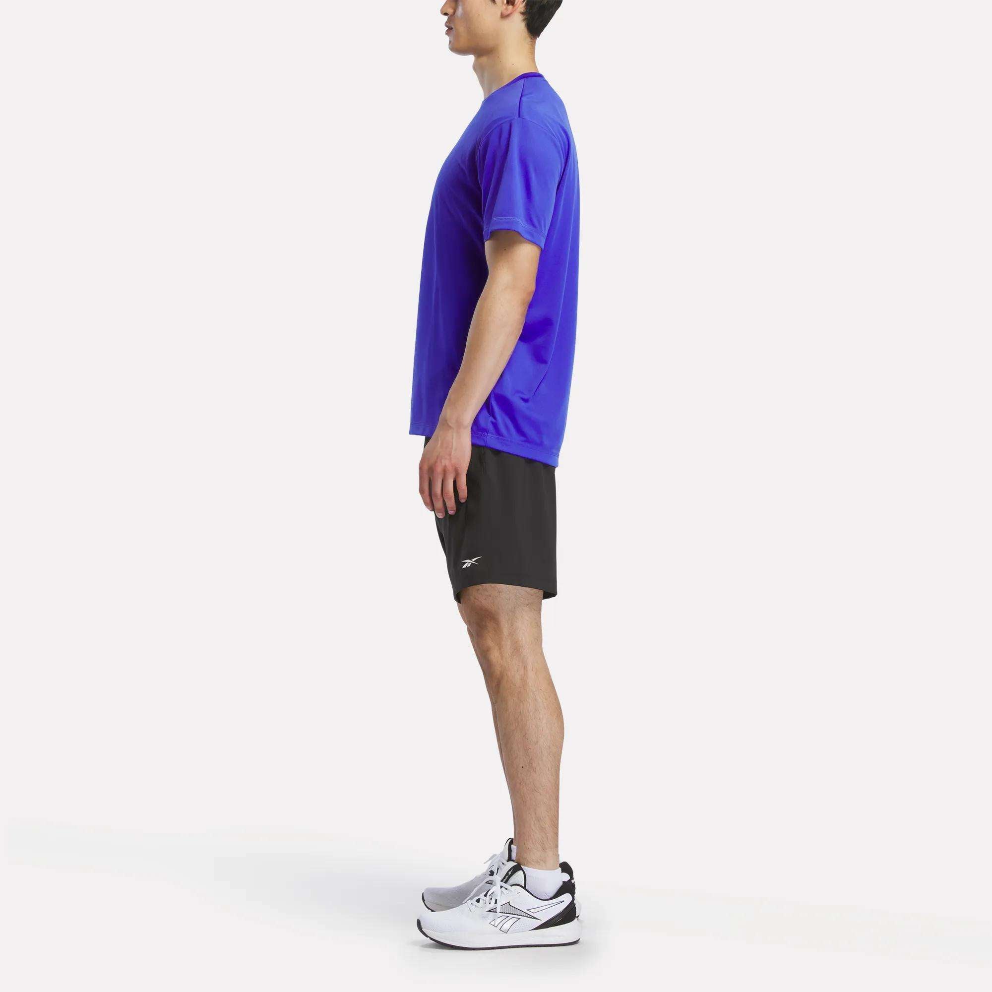 A man in profile wears a Reebok Men's Short Sleeve Tech Training T-Shirt, sweat-wicking black athletic shorts, and white sneakers, posed against a plain white background.
