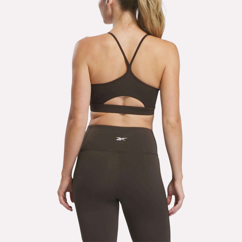 A woman wearing the Reebok Women’s Strappy Sports Bra, featuring a stylish back cutout designed for medium-impact workouts, and high-waisted brown leggings stands with her back to the camera against a plain white background.