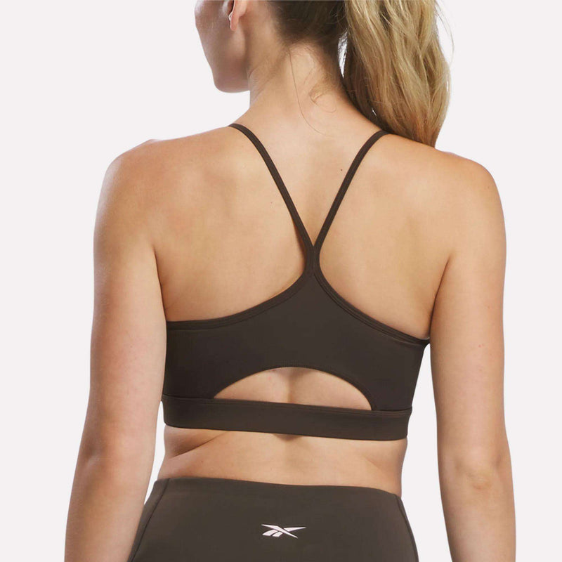 A woman with a ponytail is shown from the back, wearing the Reebok Women’s Strappy Sports Bra, which features thin straps and a small cutout designed for medium-impact workouts, paired with matching athletic leggings displaying the Reebok logo.