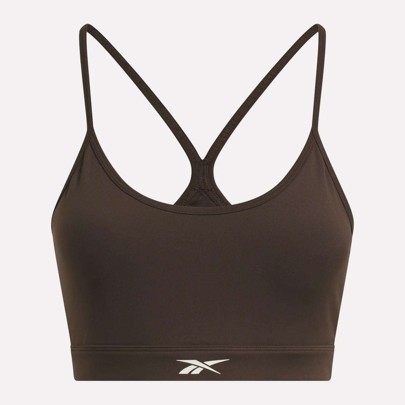 The Women’s Strappy Sports Bra by Reebok features a brown colorway with thin, crisscrossed straps in the back and a white Reebok logo centered on the front band. Designed for medium-impact workouts, this quick-drying sports bra is highlighted beautifully against a light pink background.
