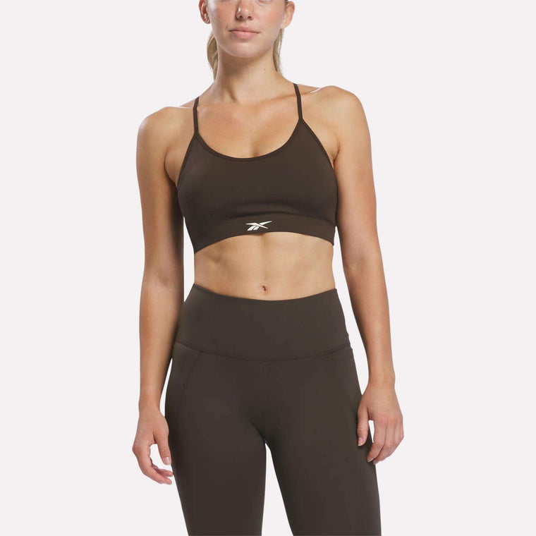 An individual stands facing forward against a plain white background, wearing the Reebok Women’s Strappy Sports Bra in espresso. Designed for medium-impact workouts, this quick-drying bra features the Reebok logo on the front. One hand rests relaxed by her side.