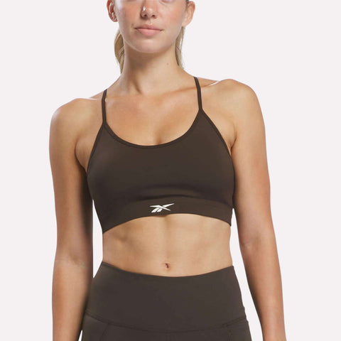 A woman stands against a plain background wearing the Reebok Women’s Strappy Sports Bra in a dark color. Designed for medium-impact workouts, this quick-drying sports bra features thin straps and is finished with a small white Reebok logo at the center.