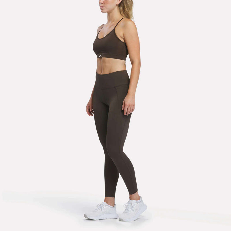 A woman stands facing left, wearing a brown Reebok Women’s Strappy Sports Bra designed for medium-impact workouts, matching high-waisted leggings, and white sneakers against a plain white background.