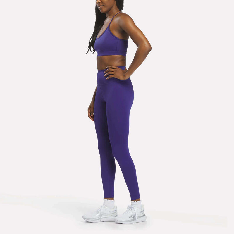 A woman stands in profile, wearing the Reebok Women's Strappy Sports Bra in purple paired with matching leggings and white athletic shoes against a plain white background—the quick-drying fabric of the sports bra makes it ideal for medium-impact workouts.