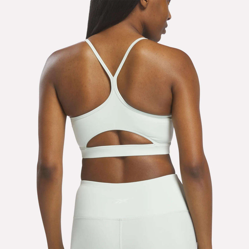 A woman stands with her back to the camera, showcasing the Reebok Women’s Strappy Sports Bra in light gray—featuring quick-drying fabric, a racerback design, and a keyhole cutout—paired with matching high-waisted leggings, making it an ideal outfit for medium-impact workouts.