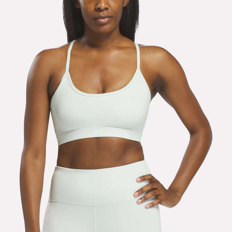 A woman stands with one hand on her hip against a plain background, wearing a light gray Reebok Women’s Strappy Sports Bra and matching high-waisted leggings—an ideal outfit for medium-impact workouts.
