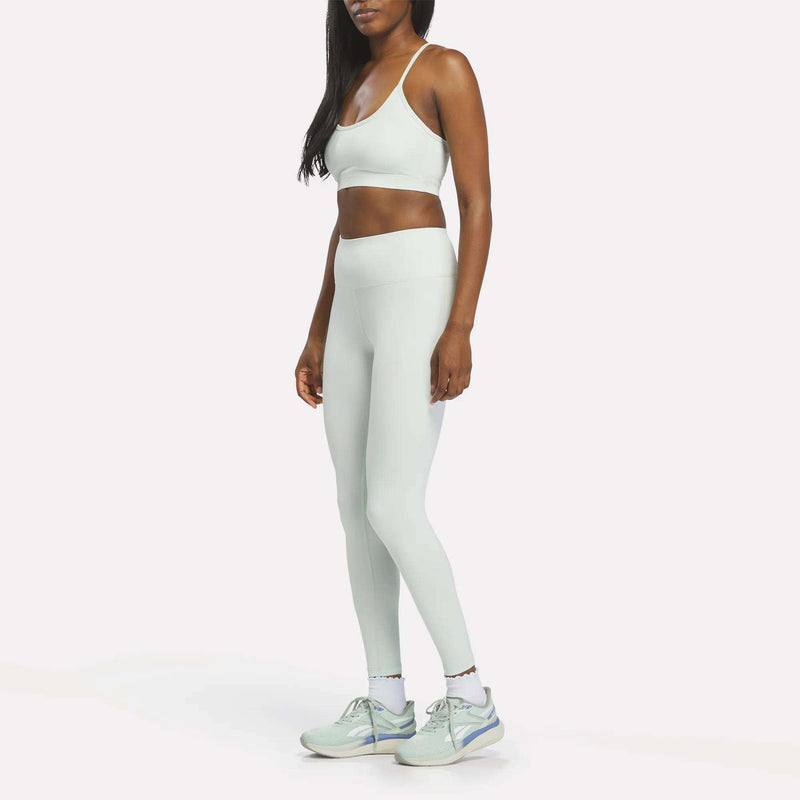 A woman stands against a plain white background, wearing the Reebok Women’s Strappy Sports Bra in light green, paired with matching high-waisted leggings and light-colored sneakers. She faces slightly to the side with her arms relaxed at her sides, ready for medium-impact workouts.