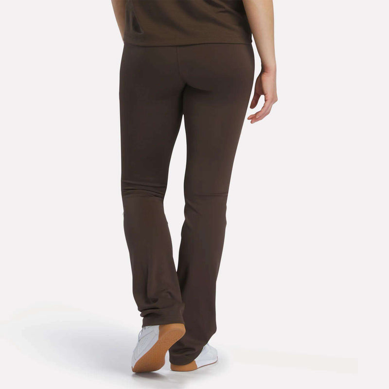 A person is seen from behind standing against a plain white background, wearing Reebok Women's High-Rise Bootcut Pants and white sneakers, with one foot slightly in front of the other.