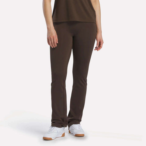 Only the lower half of a person is visible as they stand against a plain white background, wearing Reebok Women's High-Rise Bootcut Pants in dark brown and matching them with white Reebok sneakers and a coordinating top.