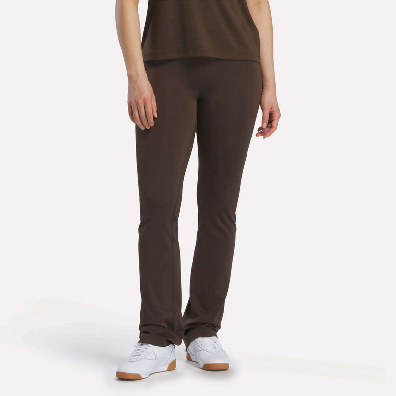 Only the lower half of a person is visible as they stand against a plain white background, wearing Reebok Women's High-Rise Bootcut Pants in dark brown and matching them with white Reebok sneakers and a coordinating top.