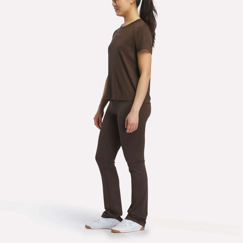 A woman stands sideways against a white background, wearing Reebok Women's High-Rise Bootcut Pants in dark brown, a matching top, and white sneakers.