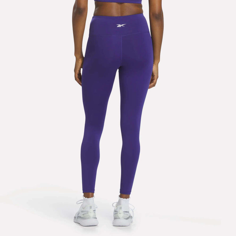 A person wearing Reebok Women's High-Rise Leggings in purple, paired with white athletic shoes, stands facing away to display the back of the high-rise leggings.