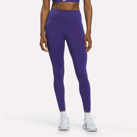 A person wearing Reebok Women's High-Rise Leggings in Bold Purple, paired with a matching sports bra and white sneakers, stands against a plain white background. Their arms are relaxed by their sides, and their face is not visible.