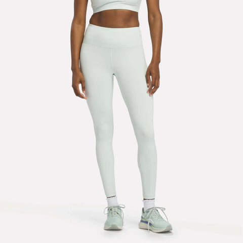 A woman dressed in Reebok Women's High-Rise Leggings in the shade Dew, paired with a matching sports bra, white socks, and mint green sneakers stands against a plain white background. Her head is out of frame.