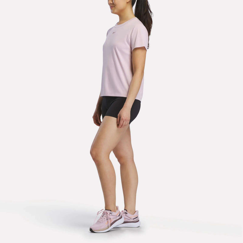 A woman stands sideways in a light pink Reebok Women's Training T-Shirt, black shorts, and pink athletic shoes. Her hair is tied back in a ponytail as she poses against a plain white background.