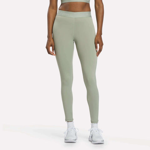 A person is shown from the waist down, standing against a plain white background, wearing Reebok Women's Ruched Training Leggings in Hazy Green, made from sweat-wicking fabric, paired with white sneakers.